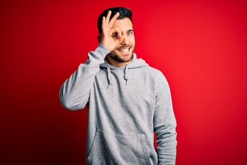 Young handsome sportsman wearing sweatshirt standing over isolated red background doing ok gesture with hand smiling, eye looking through fingers with happy face.