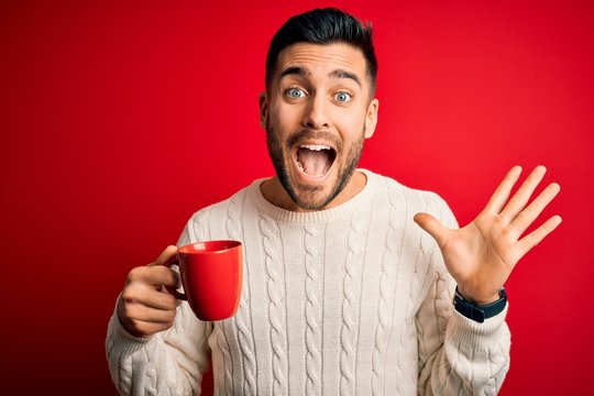 Young handsome man drinking a cup of hot coffee over red isolated background very happy and excited, winner expression celebrating victory screaming with big smile and raised hands