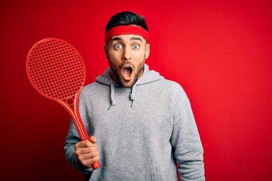 Young handsome sportsman holding tennis racket wearing sportswear over red background scared in shock with a surprise face, afraid and excited with fear expression