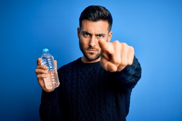 Young handsome man drinking bottle of water to refeshment over blue background pointing with finger to the camera and to you, hand sign, positive and confident gesture from the front