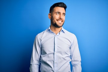 Young handsome man wearing elegant shirt standing over isolated blue background looking away to side with smile on face, natural expression. Laughing confident.