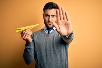 Young handsome man holding paper airplane standing over isolated yellow background with open hand...