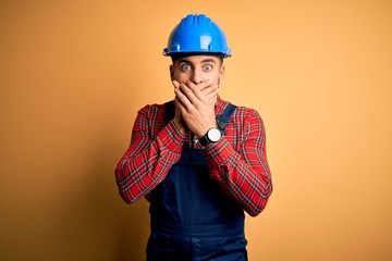 Young builder man wearing construction uniform and safety helmet over yellow background shocked covering mouth with hands for mistake. Secret concept.