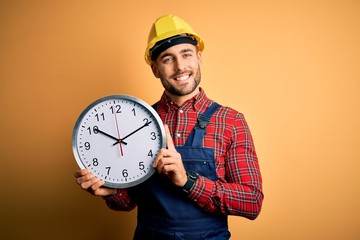 Young builder man wearing safety helmet holding big clock over yellow background with a happy face...