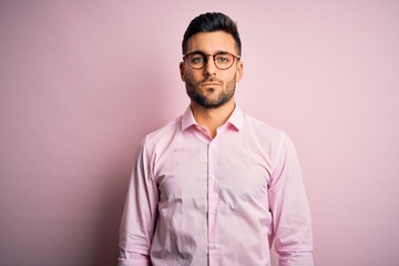 Young handsome man wearing elegant shirt and glasses standing over pink background with serious expression on face. Simple and natural looking at the camera.