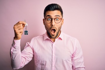 Young real estate business man holding new house keys over pink background scared in shock with a surprise face, afraid and excited with fear expression