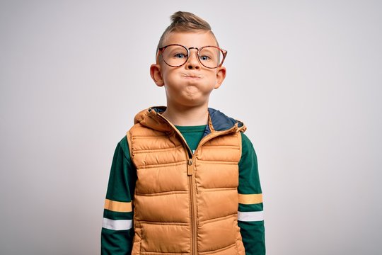 Young little caucasian kid with blue eyes wearing winter coat and smart glasses puffing cheeks with funny face. Mouth inflated with air, crazy expression.