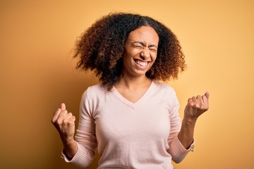 Young african american woman with afro hair wearing casual sweater over yellow background very happy and excited doing winner gesture with arms raised, smiling and screaming for success. Celebration