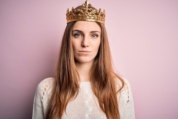 Young beautiful redhead woman wearing queen crown over isolated pink background with serious expression on face. Simple and natural looking at the camera.