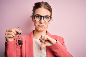 Young beautiful redhead house agent woman holding home key over pink bakcground with angry face,...