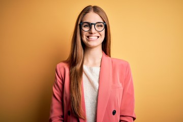 Young beautiful redhead woman wearing jacket and glasses over isolated yellow background with a happy and cool smile on face. Lucky person.