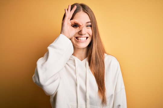 Young beautiful redhead sporty woman wearing sweatshirt over isolated yellow background doing ok gesture with hand smiling, eye looking through fingers with happy face.