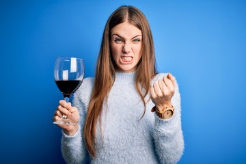 Young beautiful redhead woman drinking glass of red wine over isolated blue background annoyed and frustrated shouting with anger, crazy and yelling with raised hand, anger concept