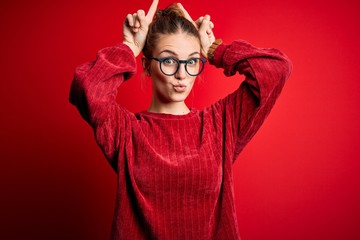 Young beautiful redhead woman wearing casual sweater over isolated red background doing funny gesture with finger over head as bull horns