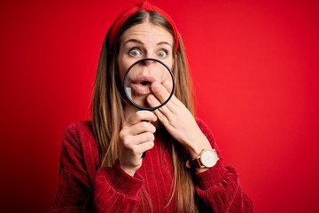 Fototapeta premium Young beautiful redhead detective woman using magnifying glass over isolated red background cover mouth with hand shocked with shame for mistake, expression of fear, scared in silence, secret concept