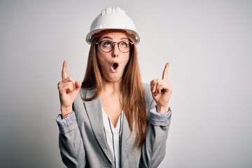 Young beautiful redhead architect woman wearing security helmet over white background amazed and...