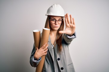 Young beautiful redhead architect woman wearing glasses and safety helmet holding blueprints with open hand doing stop sign with serious and confident expression, defense gesture