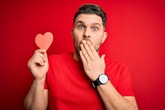 Young romantic man with blue eyes holding red heart paper shaped over red background cover mouth with hand shocked with shame for mistake, expression of fear, scared in silence, secret concept