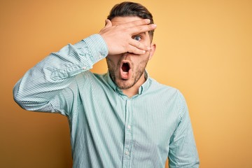 Young business man with blue eyes wearing elegant green shirt over yellow background peeking in shock covering face and eyes with hand, looking through fingers with embarrassed expression.