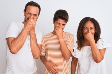 Family of three, mother, father and son standing over white isolated background smelling something stinky and disgusting, intolerable smell, holding breath with fingers on nose. Bad smells concept.