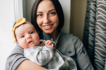 Fototapeta premium Portrait of pretty woman with black hair sits on a windowsill with her pretty little child and smiles