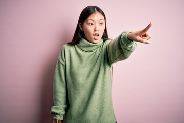 Young beautiful asian woman wearing green winter sweater over pink solated background Pointing with finger surprised ahead, open mouth amazed expression, something on the front