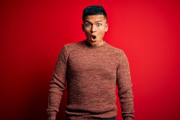 Young handsome latin man wearing casual sweater standing over red background afraid and shocked with surprise expression, fear and excited face.