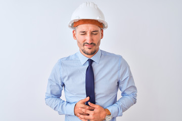Young business man wearing contractor safety helmet over isolated background with hand on stomach because indigestion, painful illness feeling unwell. Ache concept.