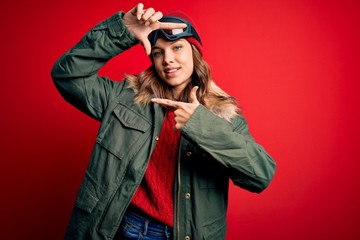 Young blonde girl wearing ski glasses and winter coat for ski weather over red background smiling making frame with hands and fingers with happy face. Creativity and photography concept.