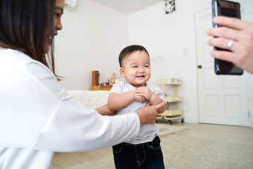 Cute excited little Asian boy enjoying funny video on mobile phone and laughing while standing in room supported by happy mom