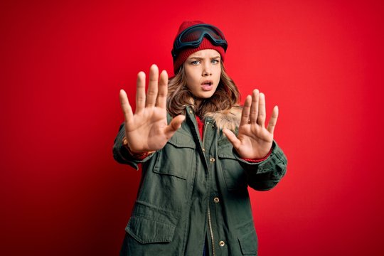 Young blonde girl wearing ski glasses and winter coat for ski weather over red background doing stop gesture with hands palms, angry and frustration expression