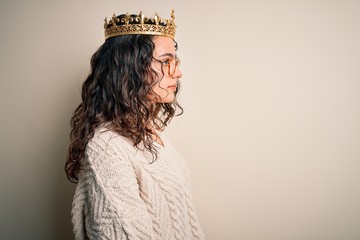 Young beautiful woman with curly hair wearing golden queen crown over white background looking to...