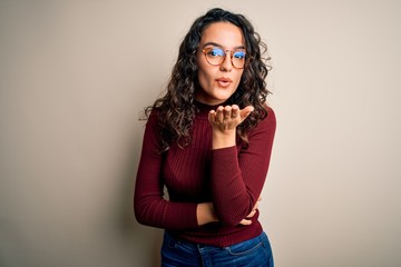 Beautiful woman with curly hair wearing casual sweater and glasses over white background looking at the camera blowing a kiss with hand on air being lovely and sexy. Love expression.