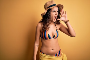 Young beautiful woman with curly hair on vacation wearing bikini and summer hat shouting and screaming loud to side with hand on mouth. Communication concept.