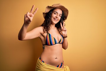 Young beautiful woman with curly hair on vacation wearing bikini and summer hat smiling looking to the camera showing fingers doing victory sign. Number two.