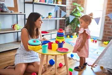 Young beautiful teacher and toddler building pyramid using hoops on the table at kindergarten