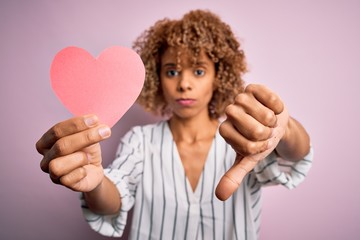 Young african american romantic woman with curly hair holding paper heart shape with angry face, negative sign showing dislike with thumbs down, rejection concept