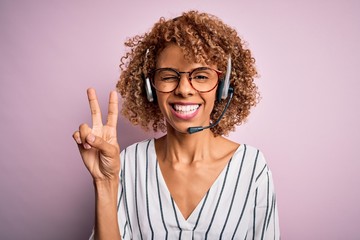 African american curly call center agent woman working using headset over pink background smiling with happy face winking at the camera doing victory sign. Number two.