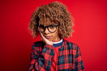 Young beautiful african american woman wearing casual shirt and glasses over red background thinking looking tired and bored with depression problems with crossed arms.