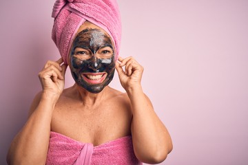 Middle age brunette woman wearing beauty black face mask over isolated pink background Smiling pulling ears with fingers, funny gesture. Audition problem