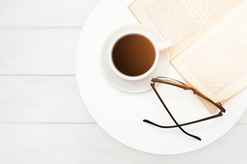 Coffee cup with an open book and glasses for reading on a white coffee table on a background light panel boards. Sunny good morning. Top view. Flat lay