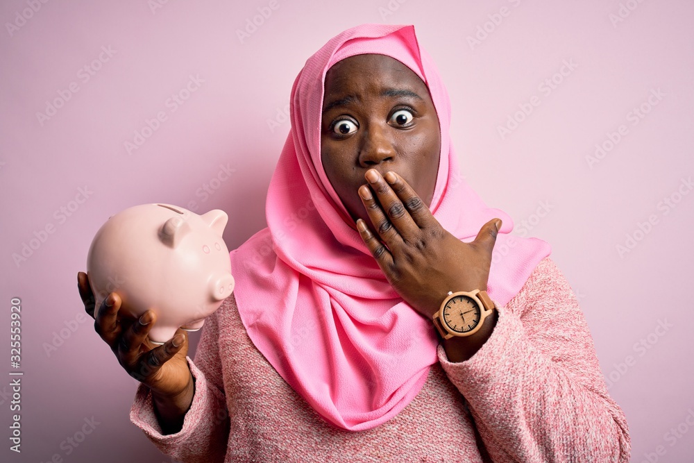 Poster Young african american plus size woman wearing muslim hijab holding pink piggy bank cover mouth with hand shocked with shame for mistake, expression of fear, scared in silence, secret concept