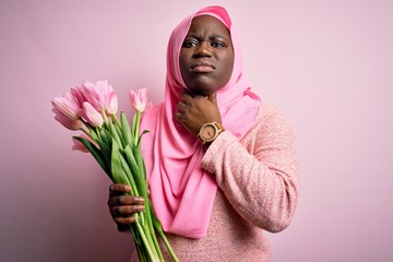 Young african american plus size woman wearing muslim hijab holding bouquet of pink tulips Touching painful neck, sore throat for flu, clod and infection