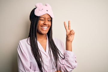Fototapeta na wymiar Young african american woman wearing pink pajama and sleep mask over isolated background smiling with happy face winking at the camera doing victory sign. Number two.