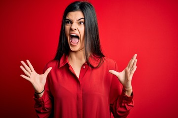 Young beautiful brunette woman wearing casual shirt standing over red background crazy and mad shouting and yelling with aggressive expression and arms raised. Frustration concept.