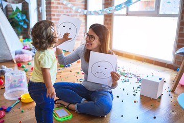 Young beautiful psicologist and toddler doing therapy using emoji emotions at kindergarten