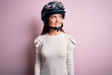 Young beautiful motorcyclist woman with blue eyes wearing moto helmet over pink background smiling looking to the side and staring away thinking.