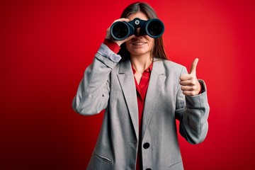 Young beautiful woman with blue eyes using binoculars over isolated red background happy with big smile doing ok sign, thumb up with fingers, excellent sign