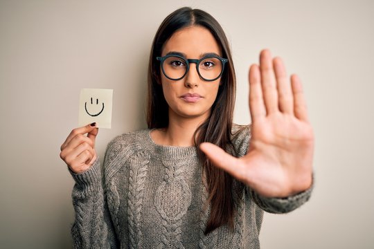 Young beautiful brunette woman wearing glasses holding paper with smile emoji with open hand doing stop sign with serious and confident expression, defense gesture