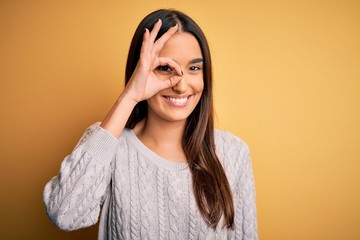 Young beautiful brunette woman wearing white casual sweater over yellow background doing ok gesture with hand smiling, eye looking through fingers with happy face.